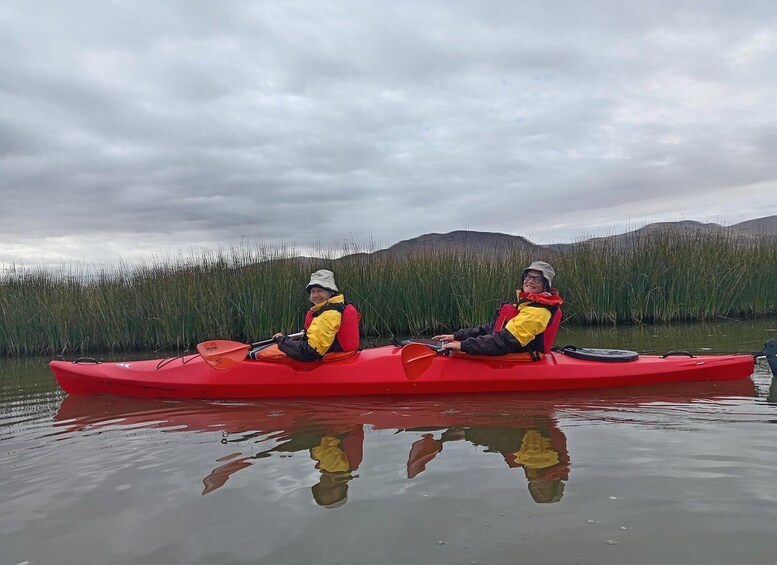 Two people kayaking in a red kayak on calm water with a grassy hill in the background. 1