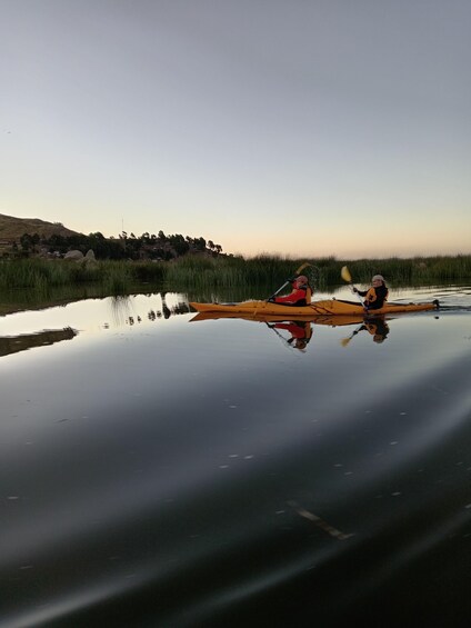 Two people kayaking on a calm body of water with reflections of the surrounding landscape. 2