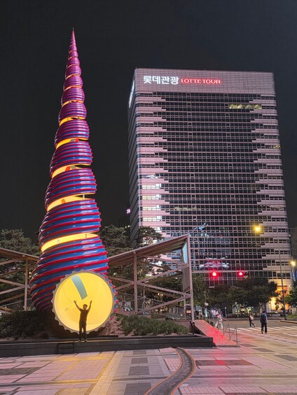 A tall, spiral structure with colorful lights, a person standing in front, and a modern building with 'LOTTE TOUR' signage in the background. 5
