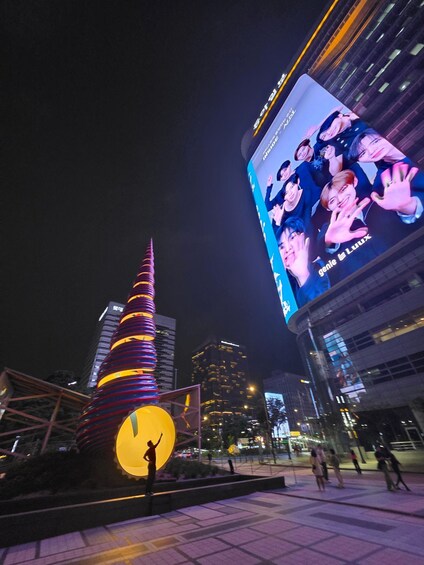 A cityscape at night with illuminated buildings, a large billboard, and a brightly lit sculpture. 2