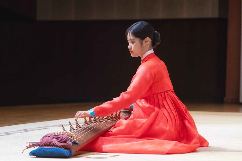 A person in a red hanbok playing a traditional Korean stringed instrument. 1