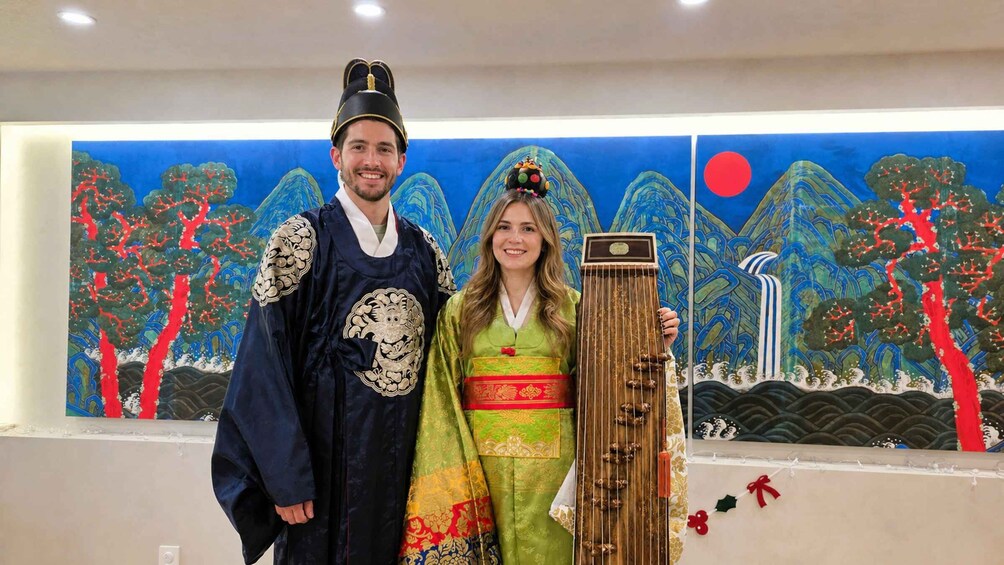 Two people in traditional attire, a man and a woman, standing in front of a mural with a mountainous landscape. 4