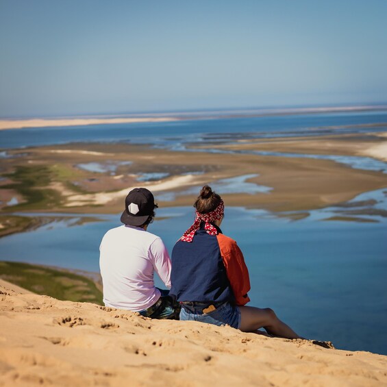 Deux personnes sont assises sur une colline de sable, d’où elles dominent un paysage côtier avec de l’eau et une terre ferme à l’horizon. 4