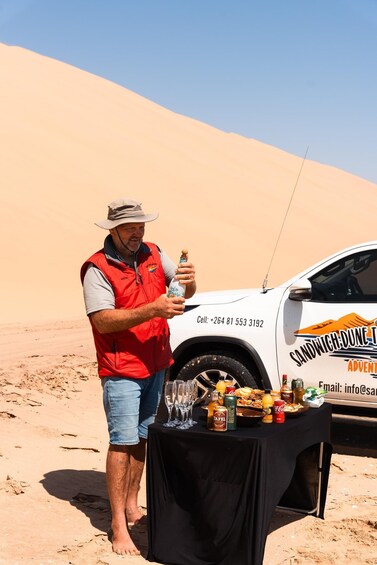 Un homme se trouve dans un paysage désertique, à proximité d’un véhicule portant l’inscription « SANDWICH-DUNES ADVENTURE », et d’une table où sont disposées des boissons et des collations. 3
