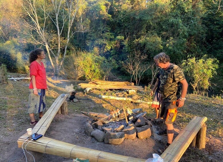 Two people near a campfire in a forested area with a makeshift wooden structure. 4