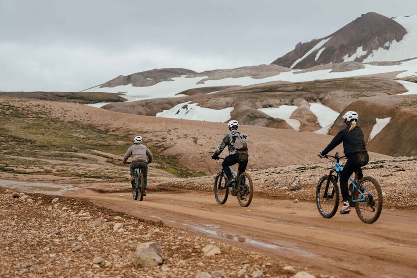 Three cyclists on a dirt road with snow patches and a mountainous backdrop. 1