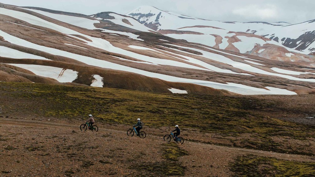 Three cyclists on a dirt trail with snow patches and a mountainous backdrop. 2