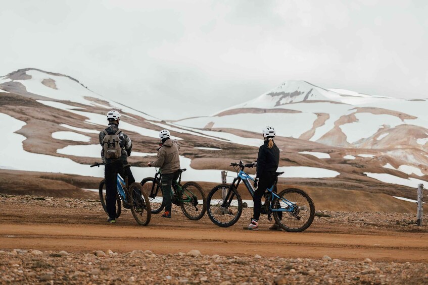 Three people with mountain bikes on a dirt road with snow patches and a mountain in the background. 4