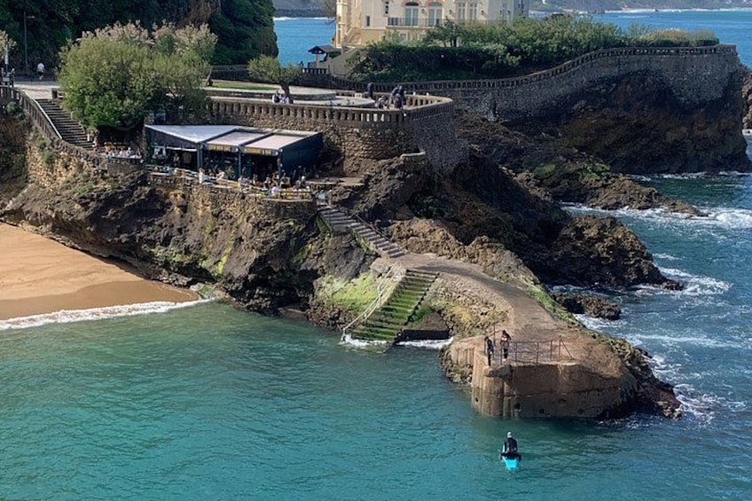A coastal area with a stone staircase leading down to the beach, a small boat in the water, and a building with a balcony overlooking the sea. 2