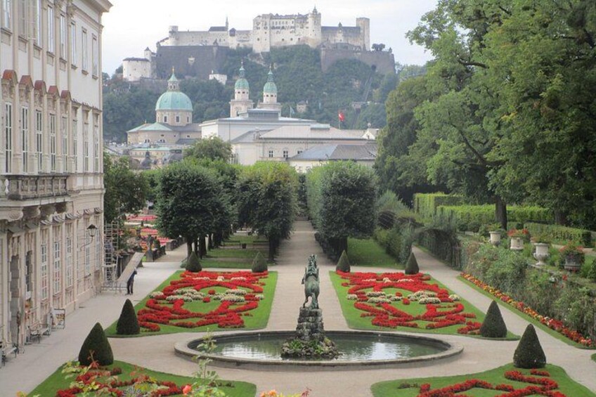 A formal garden with symmetrical flower beds, a central fountain, and a castle in the background. 1