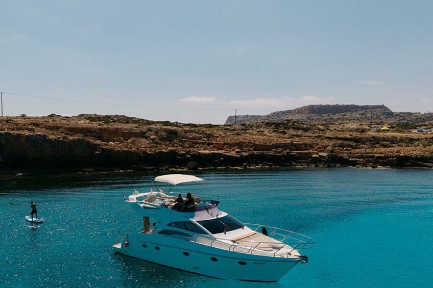 En vit yacht ligger ankrad i kristallklart blått vatten, med en klippig kustlinje i bakgrunden. 1