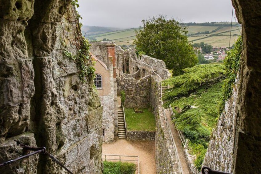A stone structure with a staircase leading to a building, surrounded by greenery and a distant village. 5