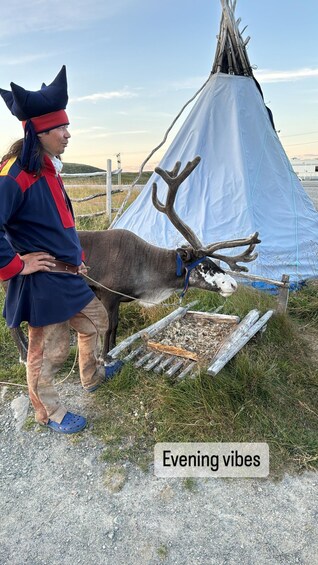 A person in traditional attire standing next to a reindeer and a teepee in a grassy outdoor setting. 5
