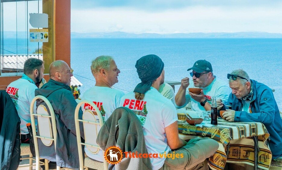 A group of people sitting at a table by the sea, wearing Peru-themed shirts, with a clear view of the ocean and distant mountains. 3