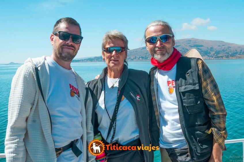 Three people standing on a boat with a clear blue sky and a mountainous landscape in the background. 1