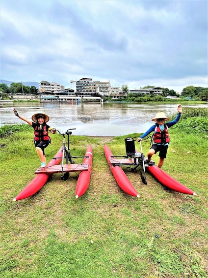 Two people standing next to red canoes on grass by a river with buildings in the background. 5