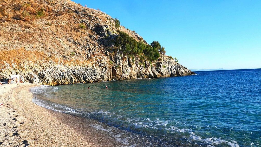 A rocky coastline with clear blue waters, a pebbly beach, and a cliff with sparse vegetation. 4