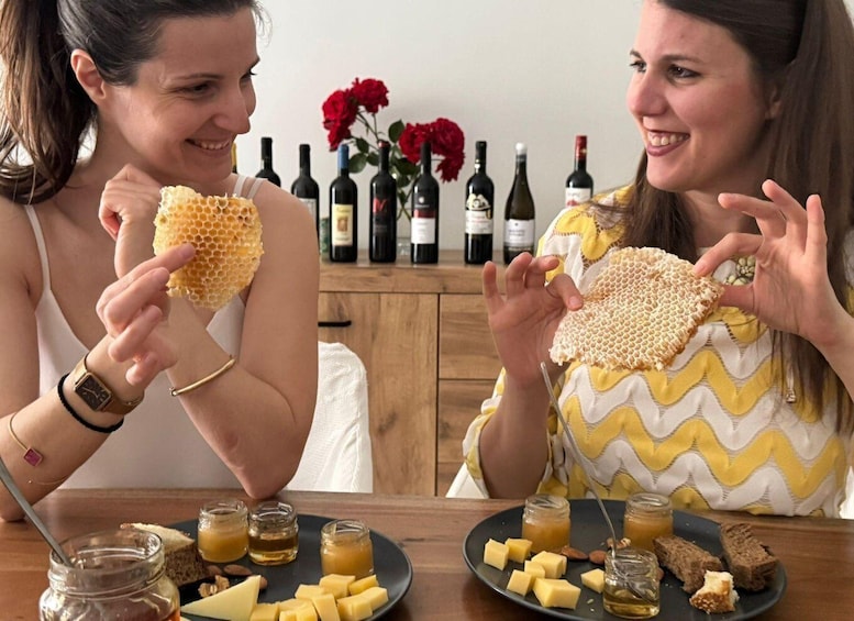 Two women holding honeycomb pieces at a table with cheese, honey, and wine bottles in the background. 4