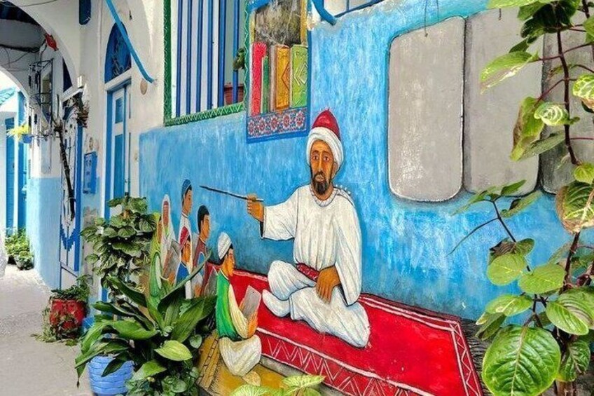 En mural forestilling af en mand i traditionelt tøj siddende på en rød måtte, med en blå bygning i baggrunden. 4