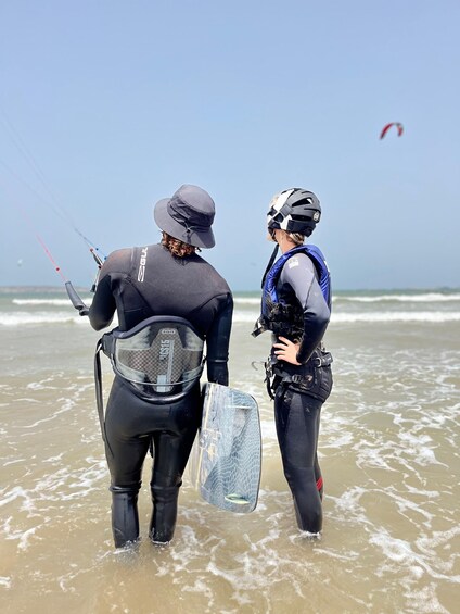 Two people in wetsuits standing in shallow water, holding a surfboard. 3