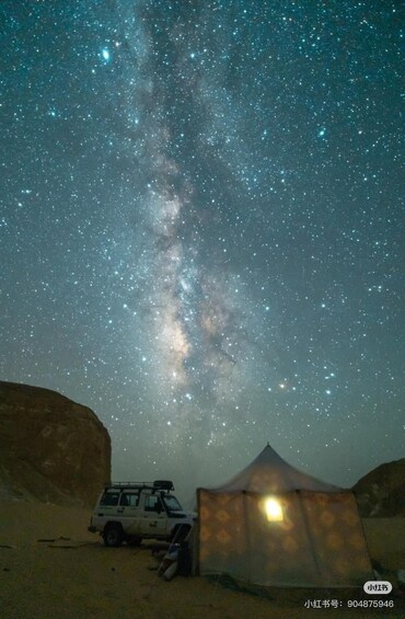 A desert night sky with stars and the Milky Way, a tent, and a vehicle. 1