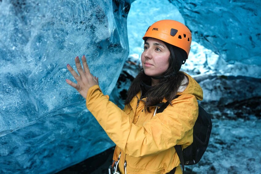 A person in a yellow jacket and orange helmet touching a blue ice wall. 1