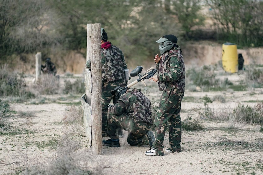 Two people in camouflage uniforms and helmets, one crouching and the other standing, in a dry, grassy field with sparse vegetation and a wooden post in the background. 3
