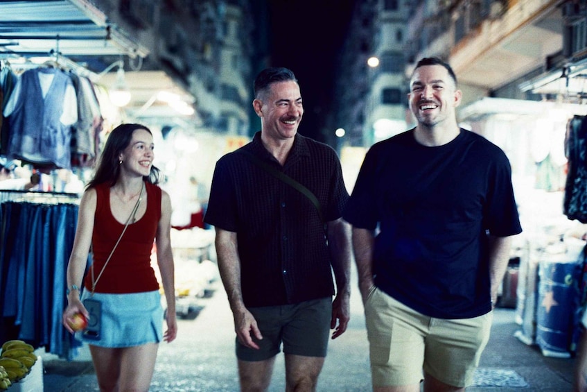 Three people walking in a market street with clothing stalls. 2