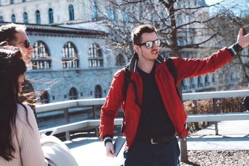 Una persona con una chaqueta roja gesticulando, con un edificio y una barandilla al fondo. 2