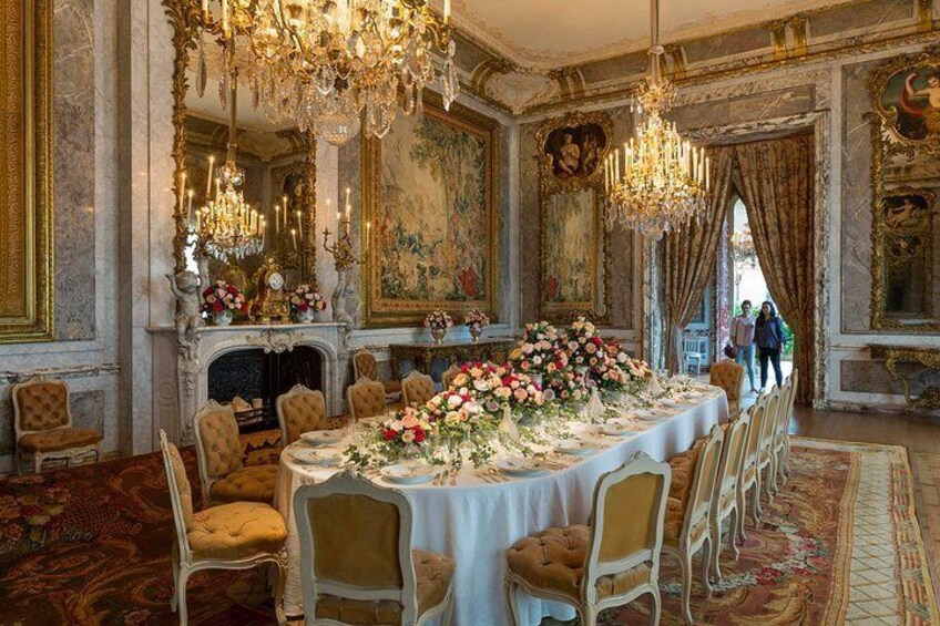 An opulent dining room with a large round table, ornate chandeliers, and classical paintings. 2