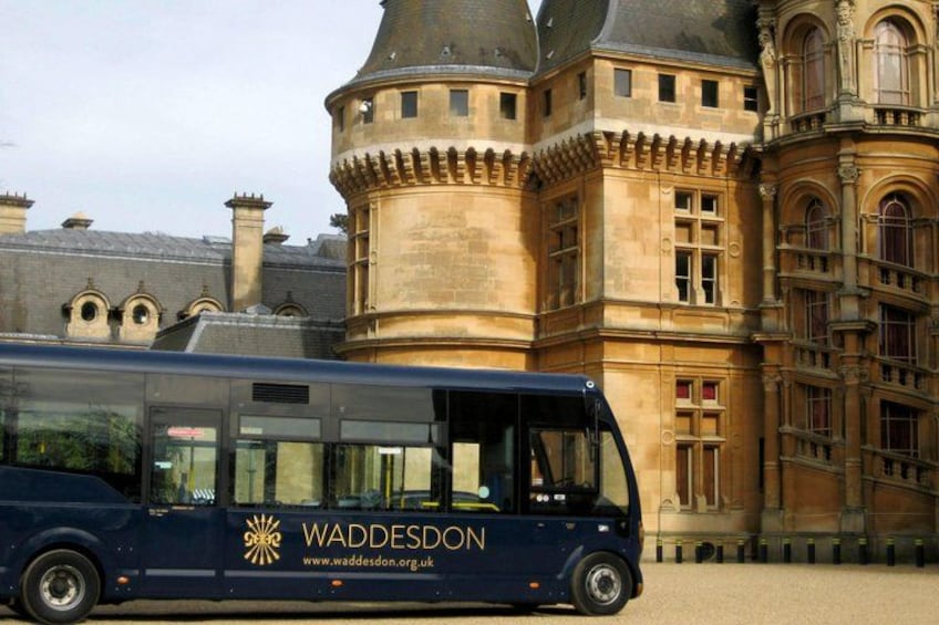 A blue bus with 'WADDESDON' and a website on its side parked in front of a historic building with a turret. 2