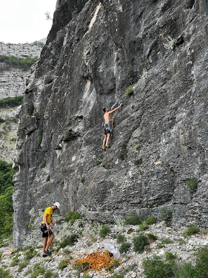 A person climbing a steep rock face with a rope, and another person assisting at the base. 3