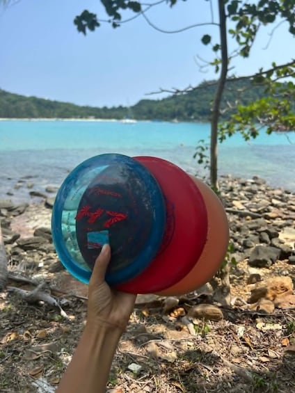 A hand holding a blue and red frisbee by a rocky lakeshore with clear water and trees in the background. 1