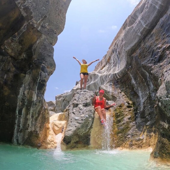 A person standing on a rock ledge with arms outstretched, another person sitting on a rock below, and a waterfall in the background. 1