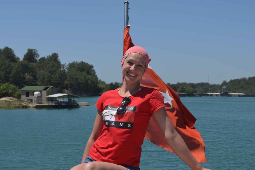 A person wearing a red shirt with a logo, sitting by a body of water with a dock and trees in the background. 4