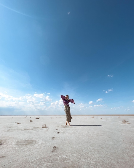 A person standing in a vast, flat, barren landscape with sparse vegetation under a clear blue sky. 5