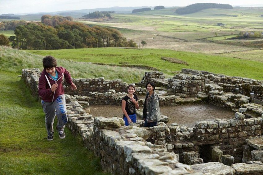 A historical stone structure with ruins, surrounded by green fields and distant hills. 1