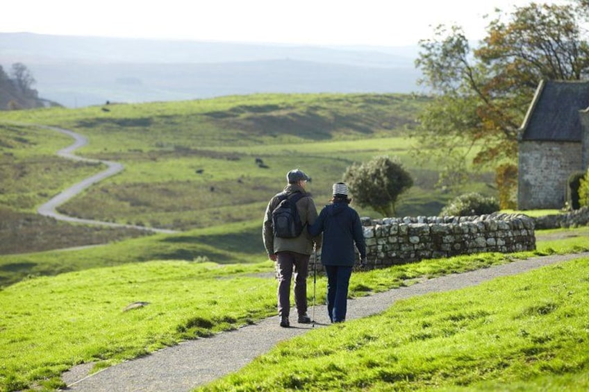 Two people walking on a path in a green landscape with a stone wall and a stone building in the background. 4