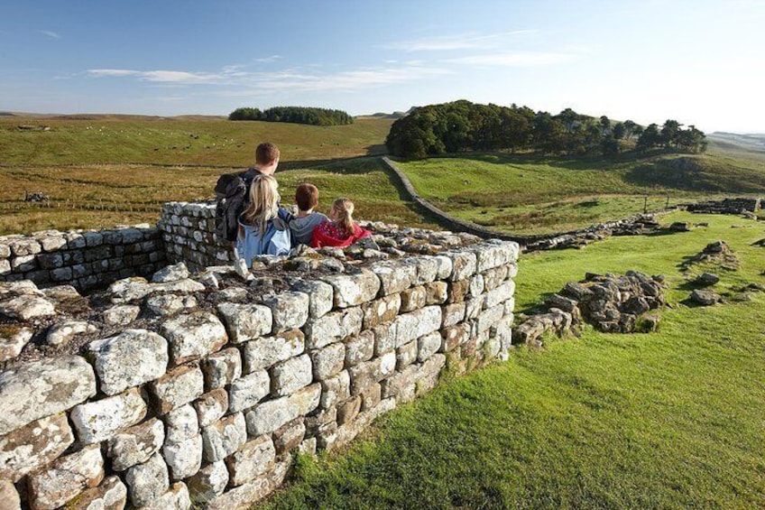 A stone wall with people standing on it, overlooking a grassy landscape with rolling hills and a distant tree line. 5
