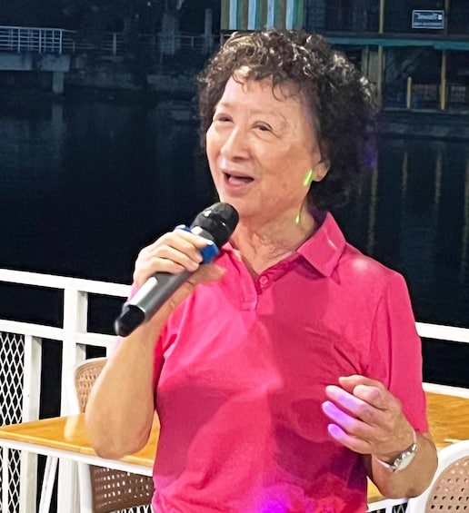 A person in a pink shirt holding a microphone, standing on a balcony with a railing and a view of a body of water. 1