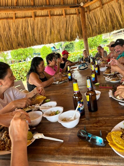 A group of people dining at an outdoor table with a thatched roof, surrounded by greenery. 5