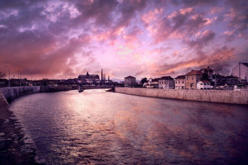 A waterfront with a stone wall, buildings, and a bridge under a colorful sky. 5