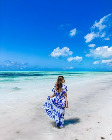 A person in a blue floral dress walking on a sandy beach with clear turquoise water and a blue sky. 3
