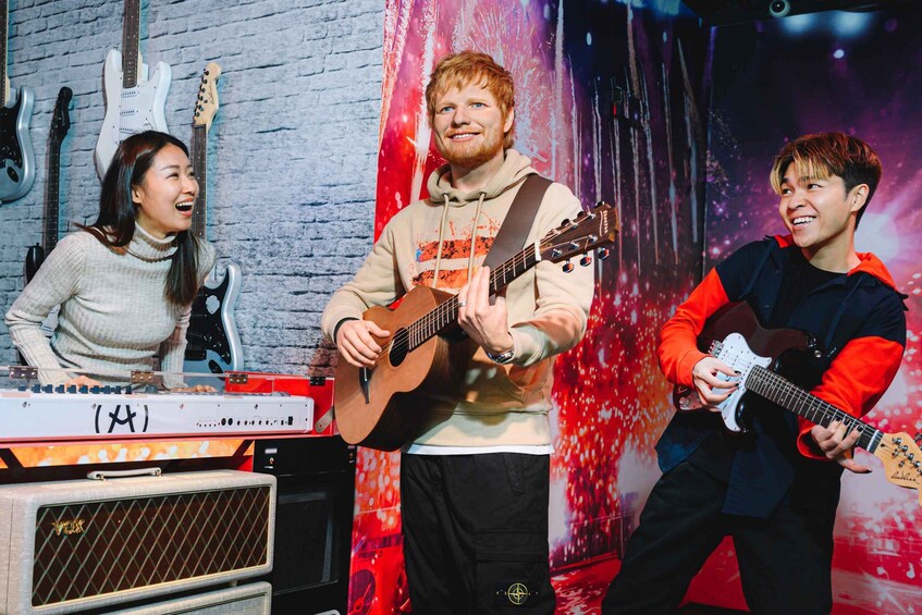 Three people playing guitars in a room with a brick wall and musical instruments. 5