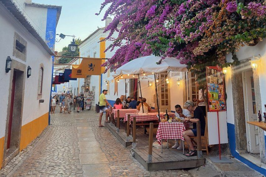 A cobblestone street with outdoor seating at a café, surrounded by buildings with colorful facades and purple flowering vines. 4