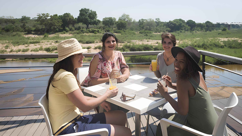 Four people sitting at a table on a deck by a river, with trees and a bridge in the background. 5