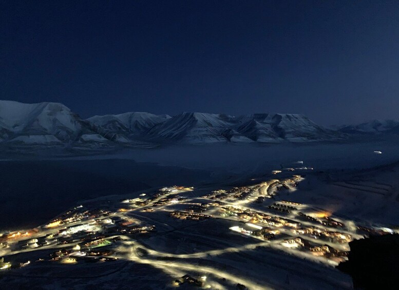 A nighttime aerial view of a snow-covered town with illuminated streets and mountains in the background. 4