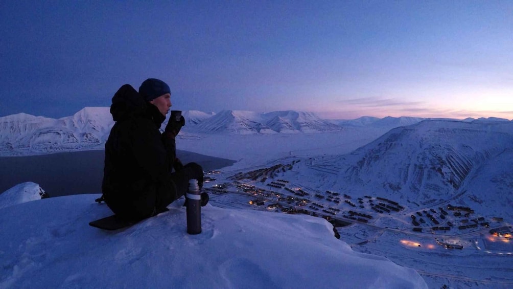 Two people sitting on a snowy peak overlooking a town with snow-covered mountains in the background. 1