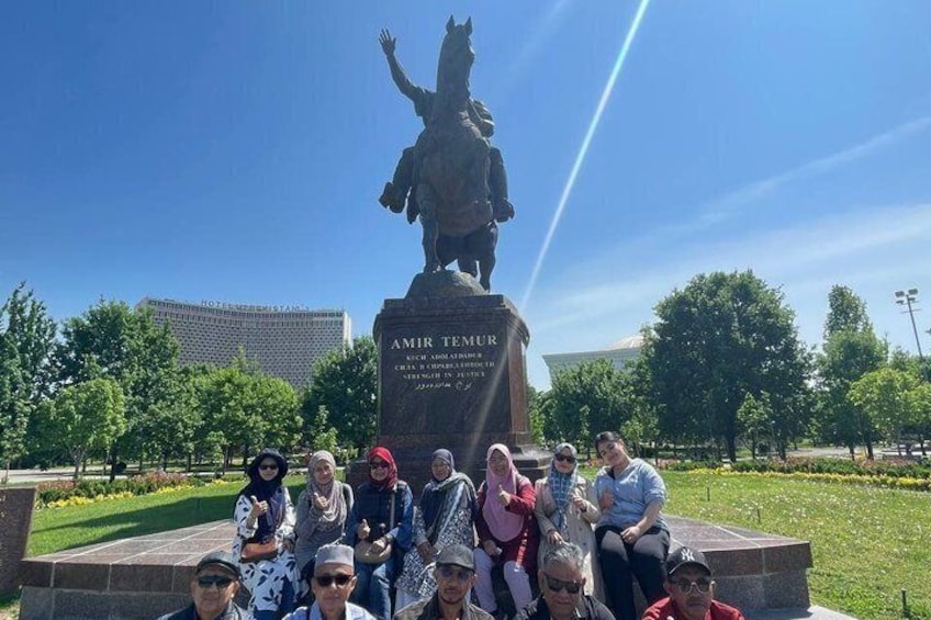 Un groupe de personnes posant devant une statue d’une figure historique, avec un ciel dégagé en arrière-plan. 1