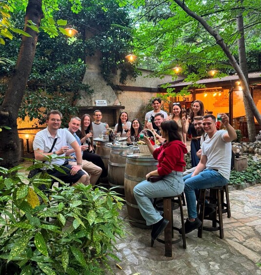 A group of people seated around barrels in an outdoor setting with greenery and a building in the background. 3